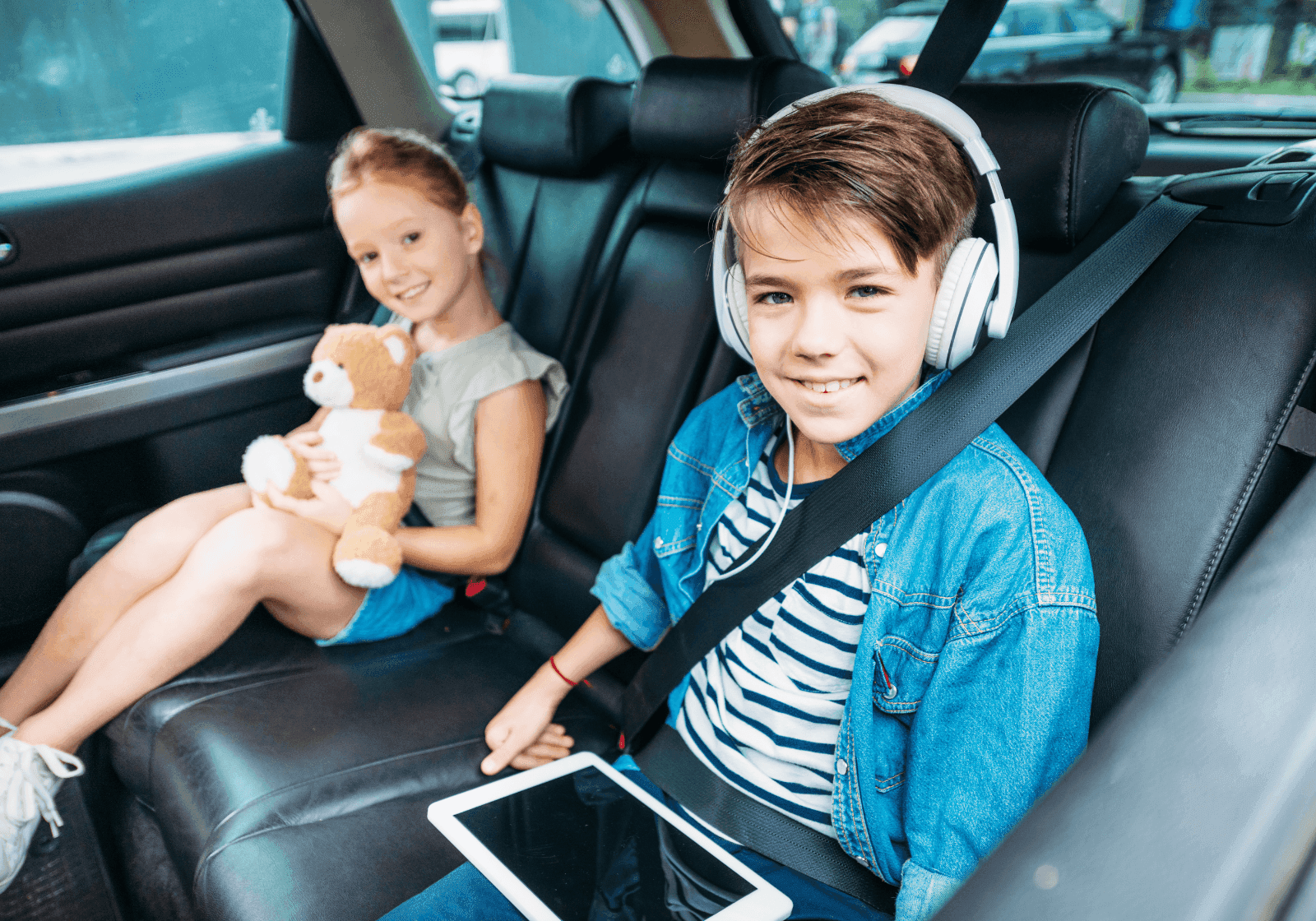 To børn på bagsædet i en bil. Den ene holder en bamse. Den anden holder en tablet og har høretelefon på