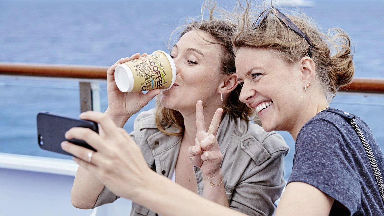 A woman sipping coffee on the ferry deck while another woman takes a selfie of them